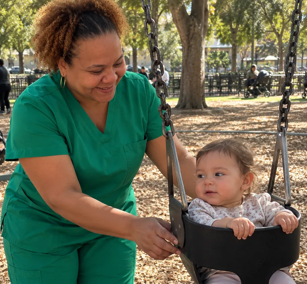 Arlene in green scrubs pushing a smiling baby on a park swing
